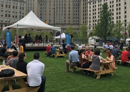 People sit at picnic benches in front of a stage where panelists are speaking